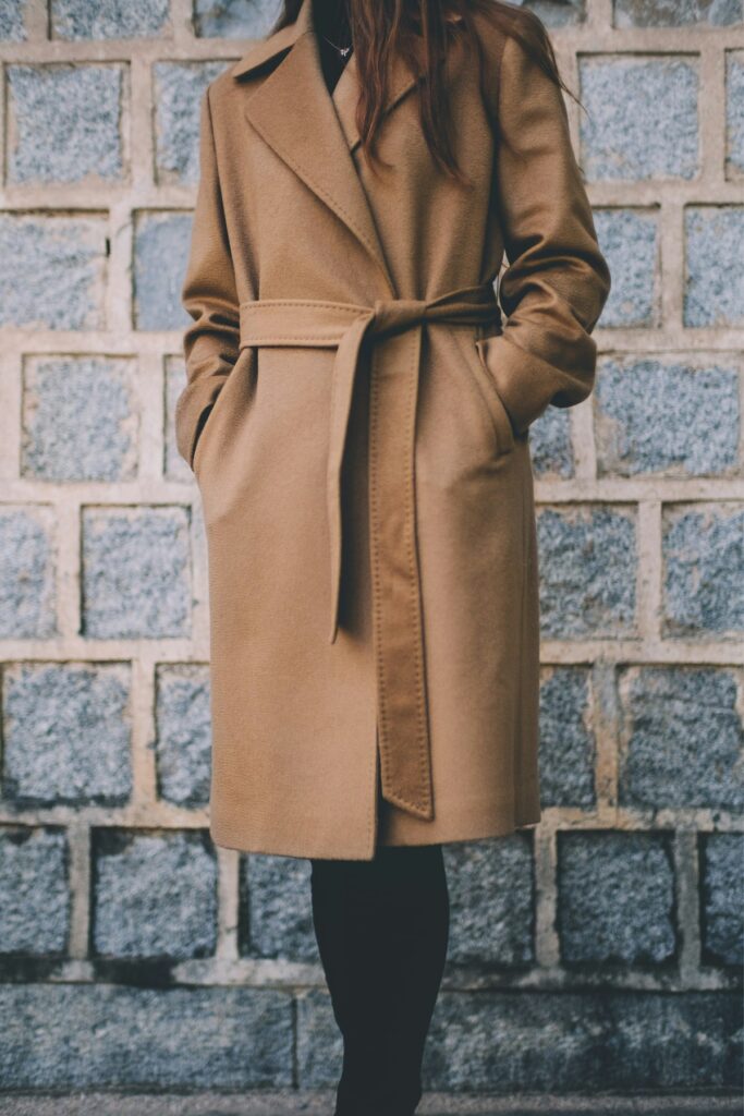 A woman in a single-breasted brown coat standing against a gray brick wall