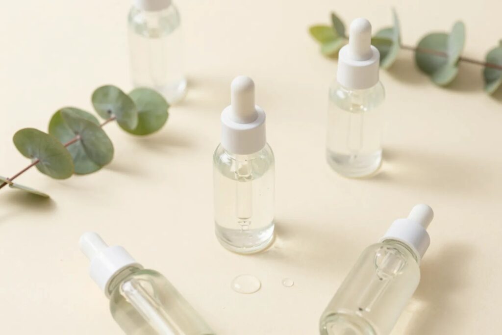Clear glass hyaluronic acid serum dropper bottles on a cream background with eucalyptus and water droplets