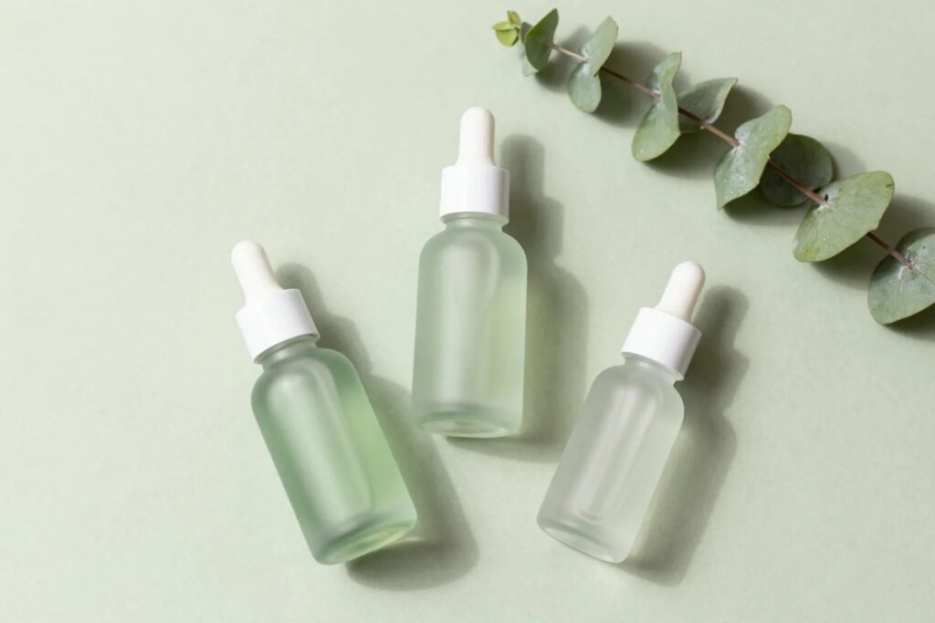 Three frosted-glass niacinamide serum dropper bottles on a sage green background with eucalyptus