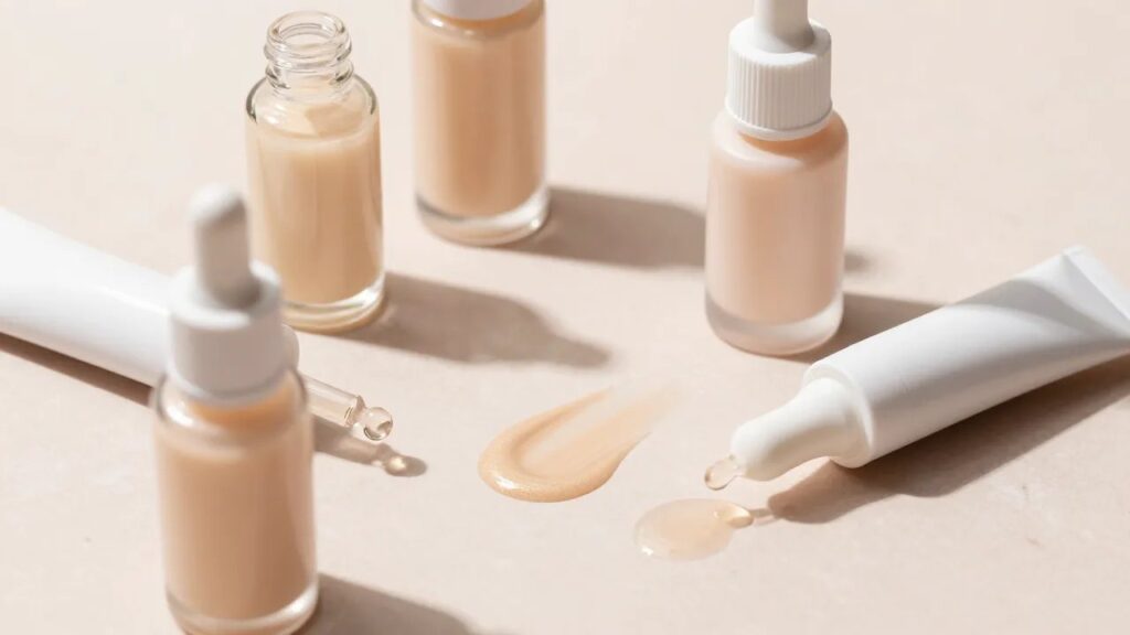 Glass dropper bottles and tubes of luminous glow filter and skin tint with a swatch on a soft beige surface